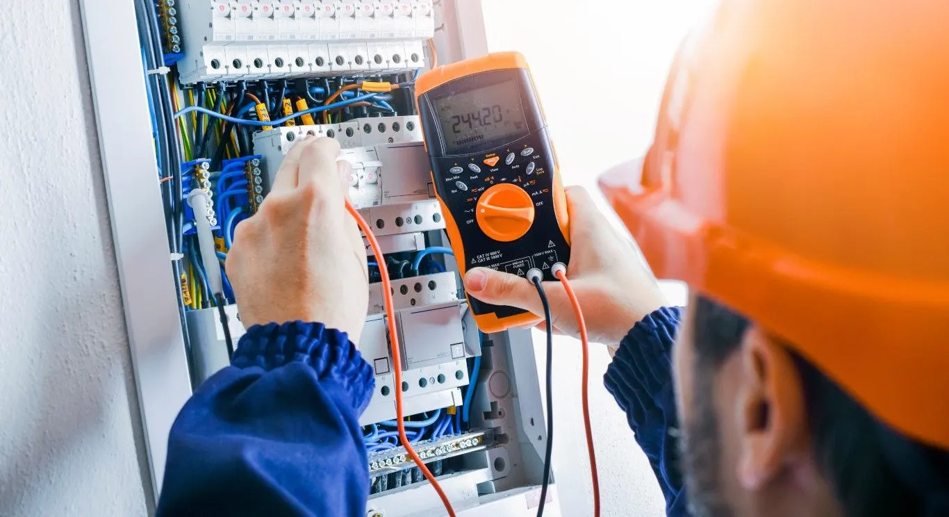 Technician testing an electrical control panel with a multimeter, wearing an orange hard hat