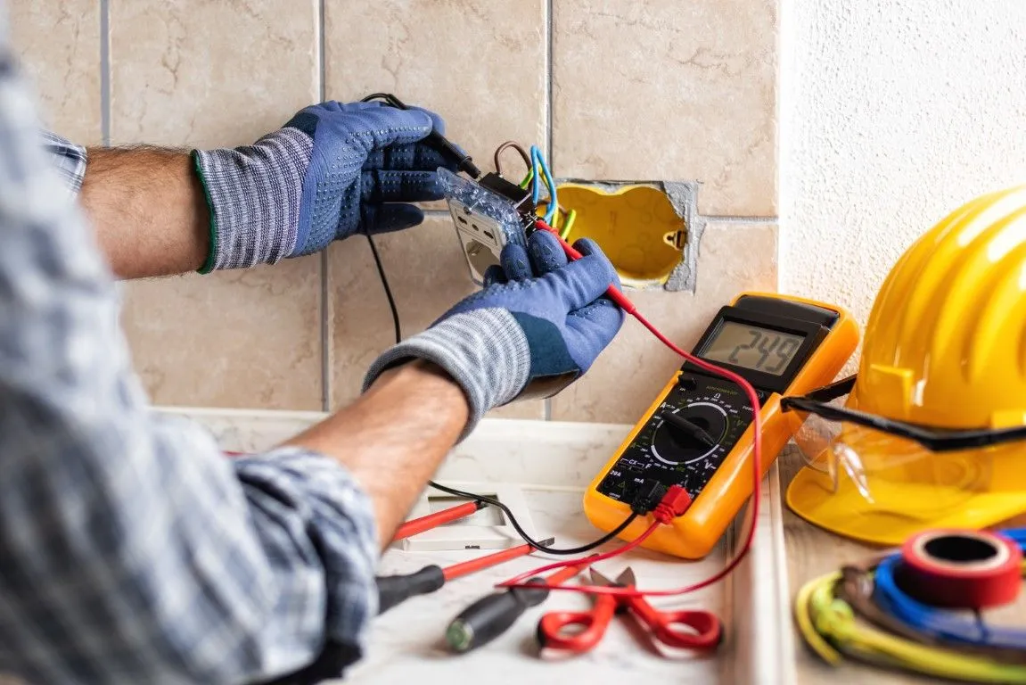 Electrician wiring a wall outlet with tools and a multimeter on a tiled work surface